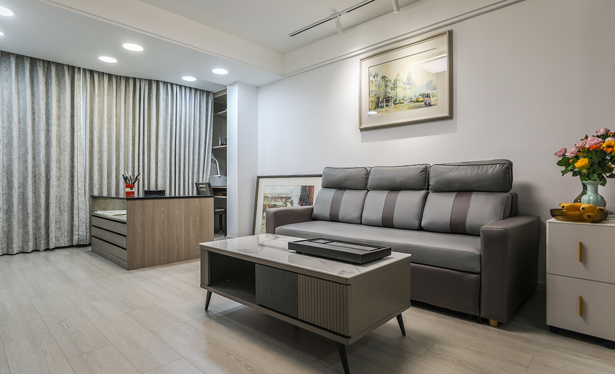 客廳是客戶平時休閑和繪畫創作的場所，要求寬闊、明亮。在陽臺區域特意設置多束燈光，提供給客戶創作需要的均勻且明亮的亮度要求。藍灰色皮質沙發配白色方型茶幾加上淺木色復合地板，造型和色調上無不給人以簡潔、大氣、時尚的感覺。