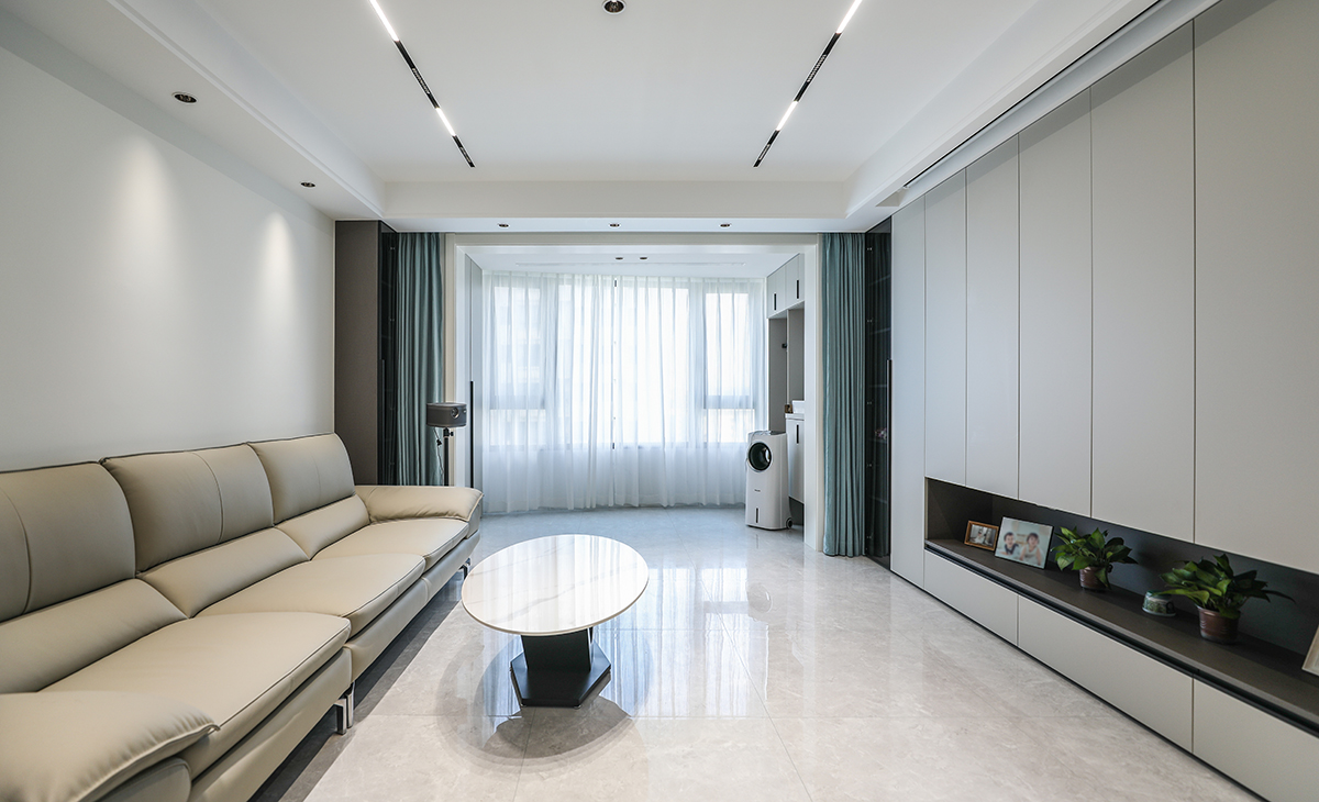 客廳開間很大，滿墻電視柜造型典雅，中間鏤空處理，適當留白，美觀大氣的同時也增加了收納空間。米灰色沙發遙相呼應，整體和諧統一，將空間感拉伸的更加開闊。吊頂造型上采用滿頂無主燈吊頂。地面采用淺灰色地磚。造型和色調上簡潔，明快，給人以一種現代感十足的高雅感受。