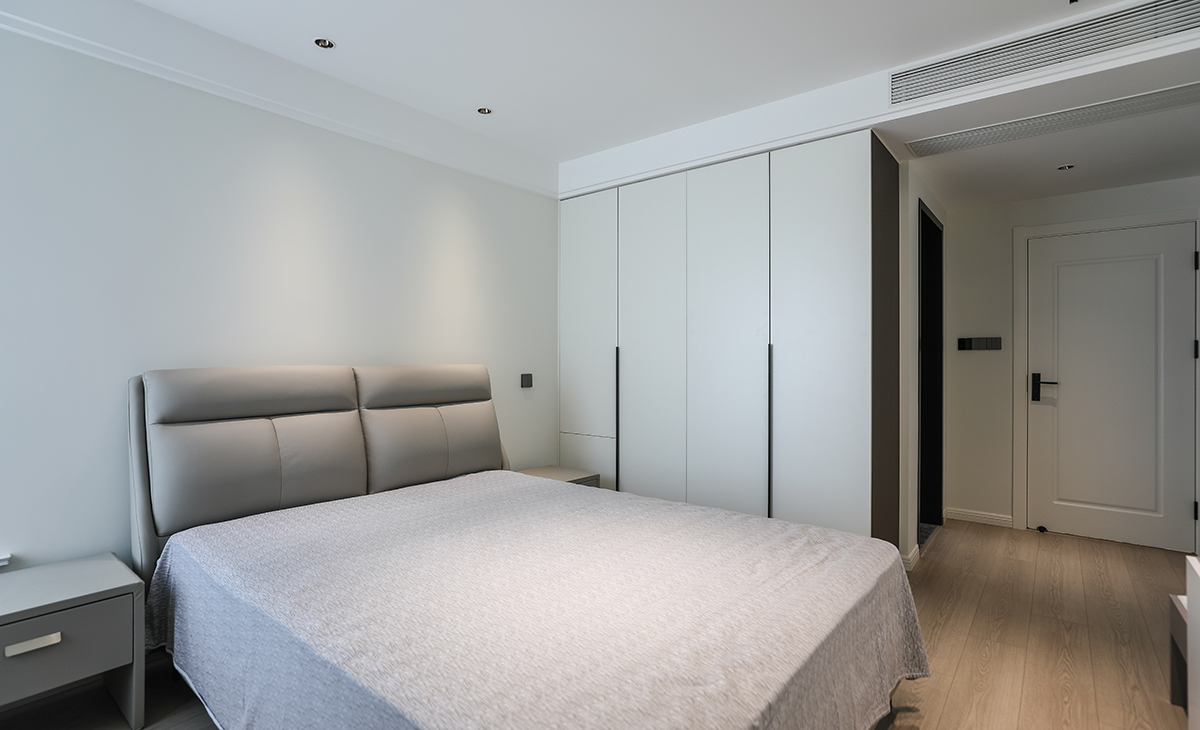 主臥地面鋪以暖灰色地板，衣柜與墻面色彩統一，相得益彰。四周雙眼皮吊頂加上中間拉平無主燈的設計，讓空間顯得尤為寬敞，灰色的床及床頭柜與整個風格進行搭配，渾然一體，整體協調，盡顯主人高端品味。