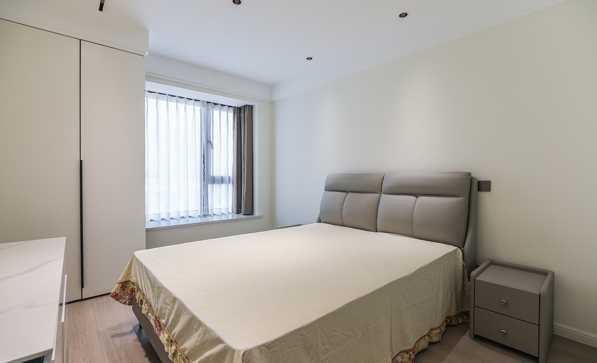 次臥地面同樣鋪以暖灰色地板，與其他房間相呼應，窗簾的色彩與床的色彩接近，體現了主人在軟裝上的極致追求。衣柜靠窗，不落俗套，讓空間布局更合理適宜，白色臺面床頭柜，整潔大方，配上吊頂里鑲嵌的筒燈，讓人有一種心曠神怡的感覺。