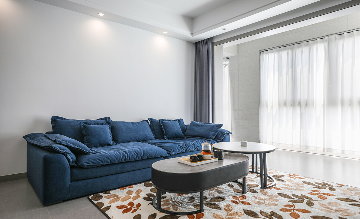 沙發背景刷淺灰色涂料，白色的沙簾，沙發背景留白設計，色調上墻面涂料和窗簾用淺色，沙發用深藍色，深淺搭配的錯落有致，既溫馨又簡潔。