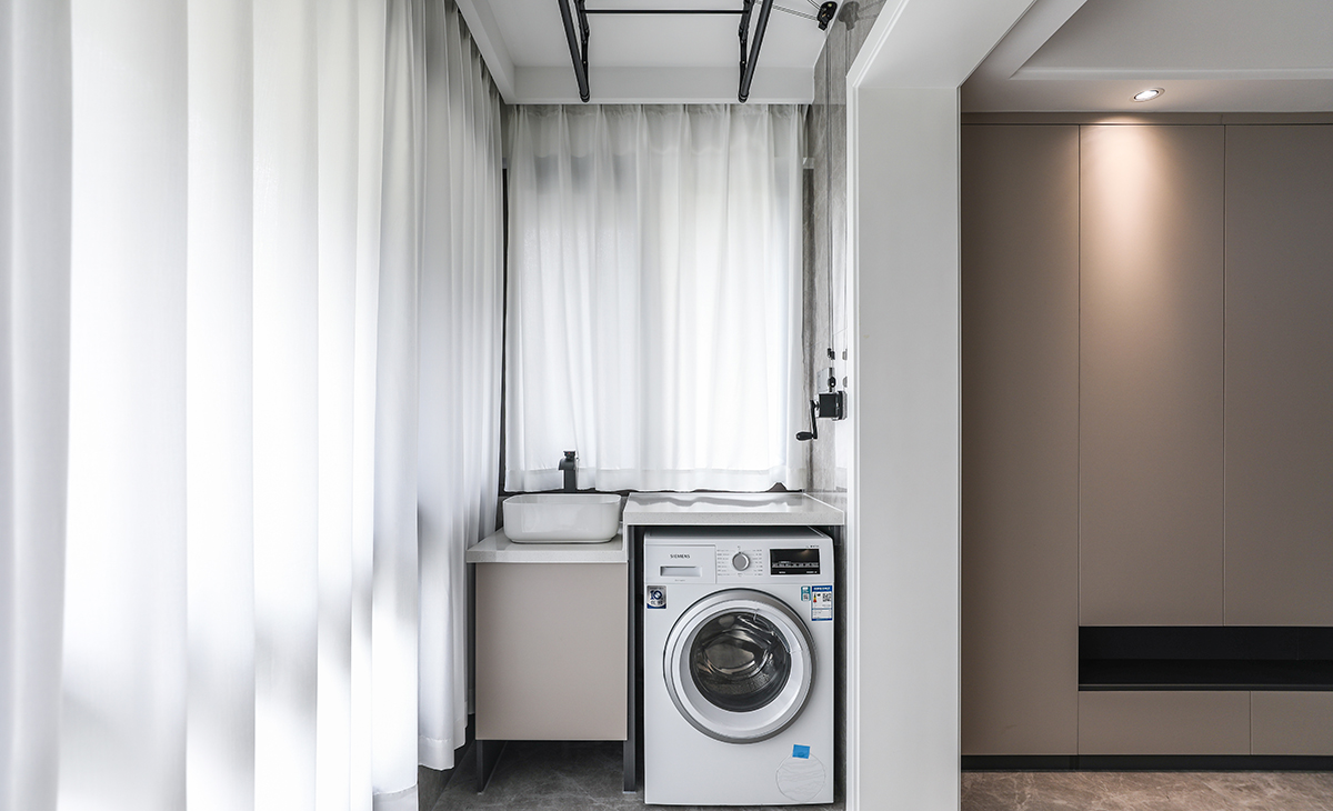 陽臺落地玻璃，洗衣柜的一面砌了矮墻，方便排水管，整體更美觀。陽臺的承重墻兩邊對應(yīng)著一面儲物柜，一面洗衣機臺盆組合柜，簡約大氣，功能合理分配。