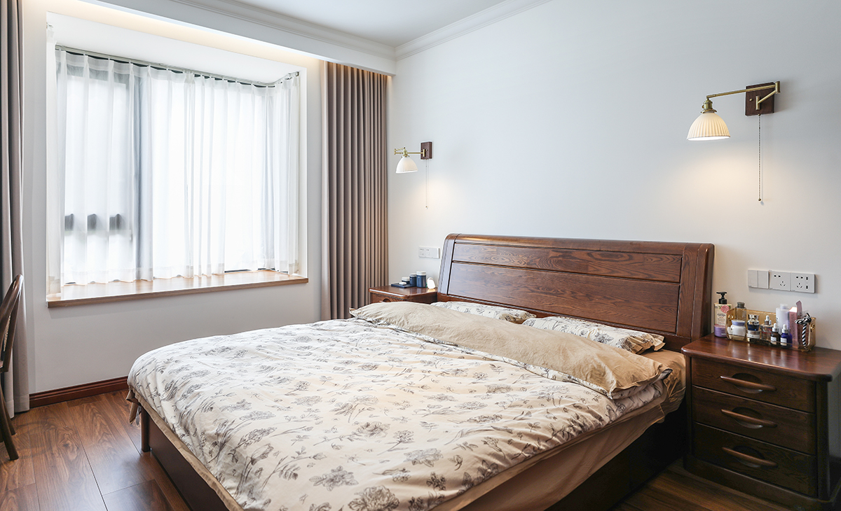 主臥床頭背景白色，地面用胡桃色，L型大飄窗讓臥室空間具有延伸感，壁燈把整個空間點綴地豐富、有內涵。