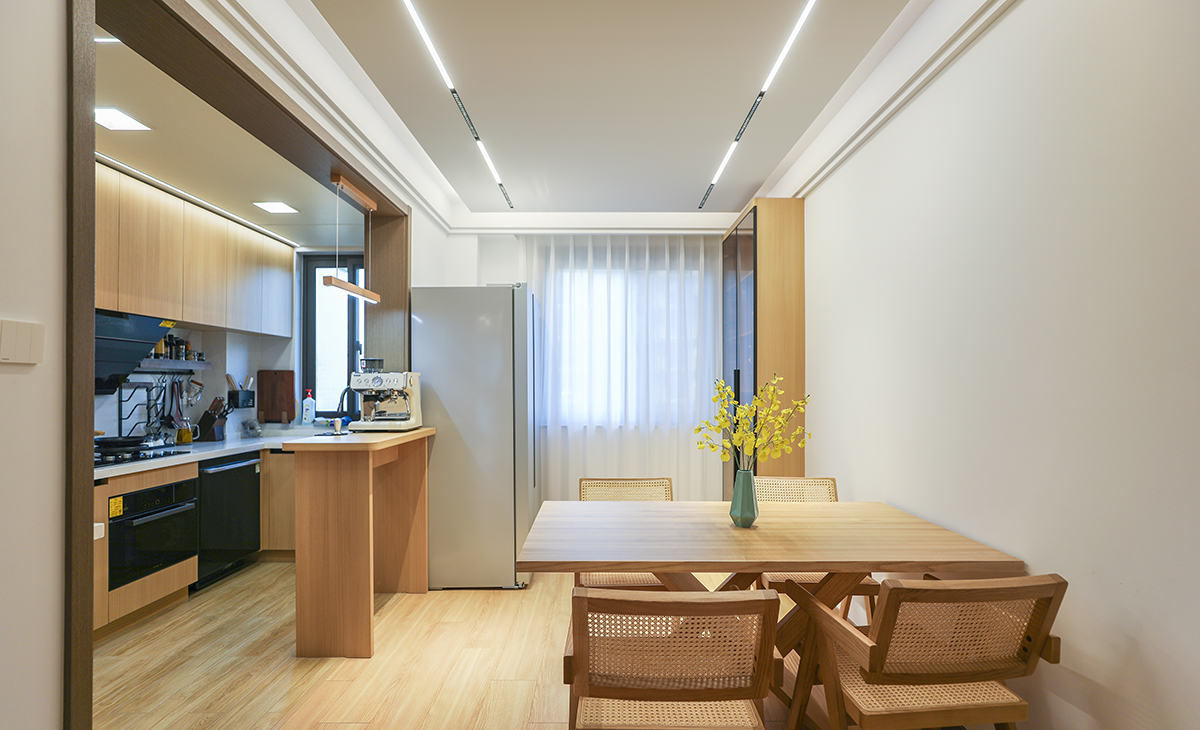 餐廳空間原本連著陽臺，把原有的陽臺敲掉，左邊放了一個冰箱，右邊放了一個玻璃柜，配上原木色的餐桌和椅子，線性燈拉長了整個空間，空間視野效果更明亮、寬敞。