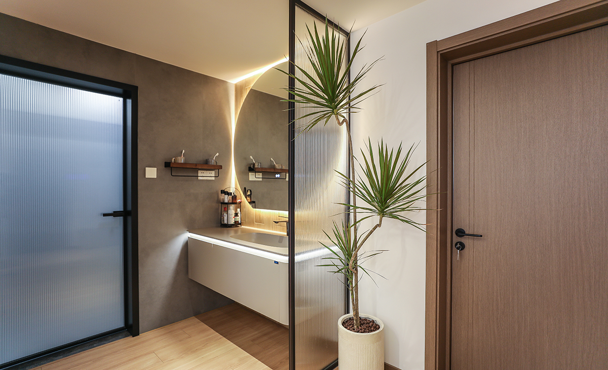 進(jìn)門口中間正好是個走道，做了個干濕分離，用現(xiàn)在流行的長虹玻璃做隔斷，透光而有私密感，墻面采用黑色水泥磚，用原木色木紋磚上墻，氛圍感一下就拉滿了，圓形的鏡子配上線性燈，整體效果圖凸出，時尚、大氣、簡單。