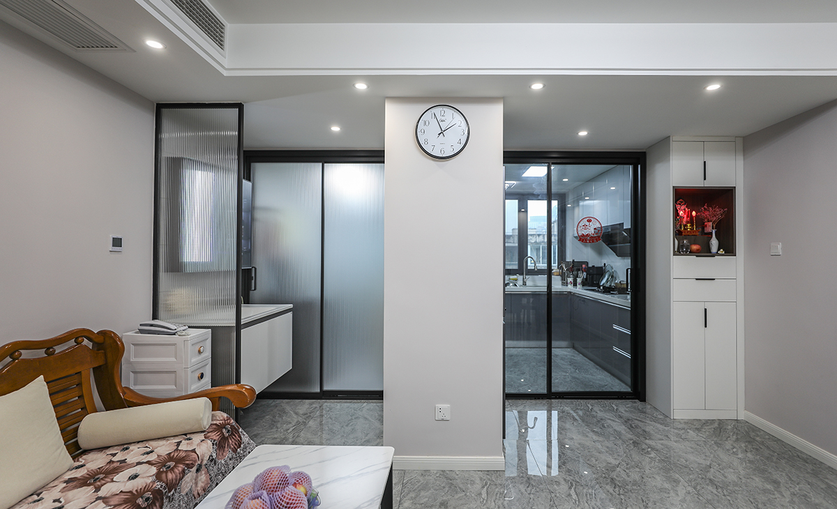 廚房和客廳的隔斷非常獨特。通過玻璃門作為隔斷，客廳內的物品和廚房的烹飪活動可以相互展現，增加了空間的通透感。沙發和桌子是客廳的基本家具，為人們提供休息和工作的空間。而那個時鐘則提醒我們珍惜時間，同時給整個空間增添了一份溫馨的氛圍。