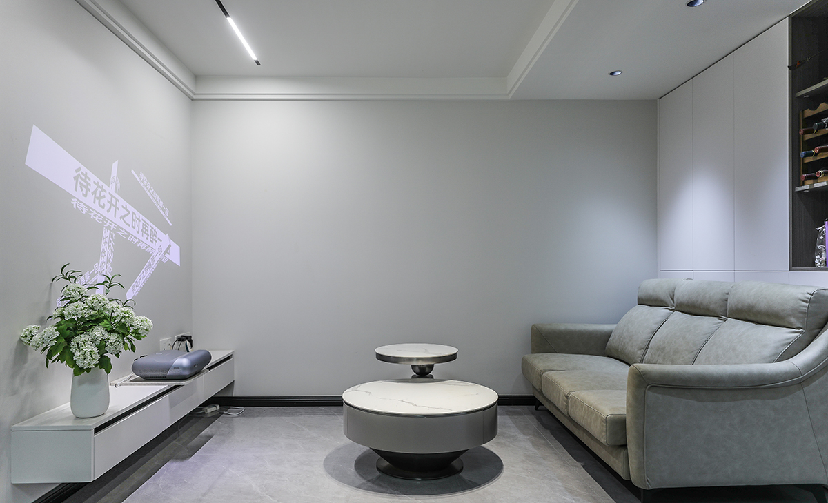  客廳開間很大，電視墻做白色乳膠漆墻面用做投影幕布墻，配合掛墻式電視柜，沙發背景白色門板配深灰開放隔顏色相互配合，明暗有神，淺灰色皮質沙發立刻檔次提高很多，放一組高低巖板茶幾柜，客廳彰顯靈動。吊頂造型上采用雙眼皮無主燈吊頂。地面采用淺灰色地磚。造型和色調上無不給人以簡潔、大氣、時尚的感覺。