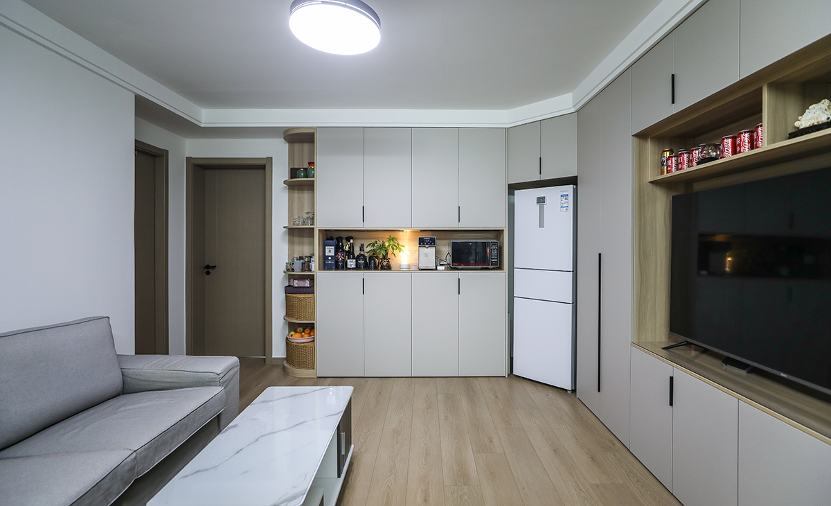  客廳開間很大，電視柜于展示柜淺色的門板和原木色木飾面相互配合，明暗有神，深灰色皮質沙發立刻檔次提高很多，客廳空間也很寬闊。吊頂造型上采用雙眼皮吊頂。地面原木色地板。造型和色調上無不給人以簡潔、舒適、溫馨的感覺。