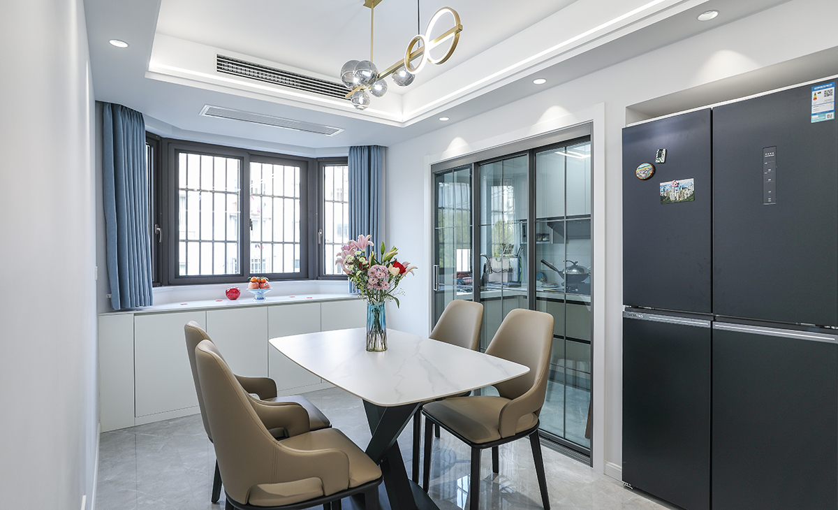 餐廳空間比較方正寬敞，又做了一個大飄窗矮柜，既實用、又有裝飾功能，冰箱為獨立位也給餐廳增加了一絲溫馨感，極窄邊框玻璃門和白色木門板相呼應，巖板餐桌把用餐氛圍調和的很溫暖，淺灰色地磚很干凈，色彩和造型上都簡潔大氣。