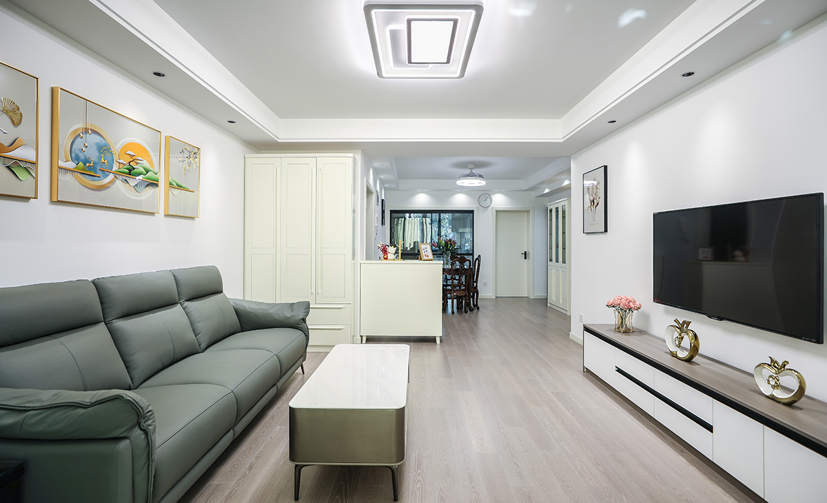  客廳面積不大，但經過設計師的手筆，讓空間緊湊不擁擠，電視墻落地電視柜，柜子色系和沙發相互配合，明暗有神，深灰色皮質沙發立刻檔次提高很多。吊頂造型上采用雙眼皮圍邊吊頂，讓造型簡介配合筒燈打造氛圍感。地面采用棕色地板。造型和色調上無不給人以簡潔、溫馨、時尚的感覺。