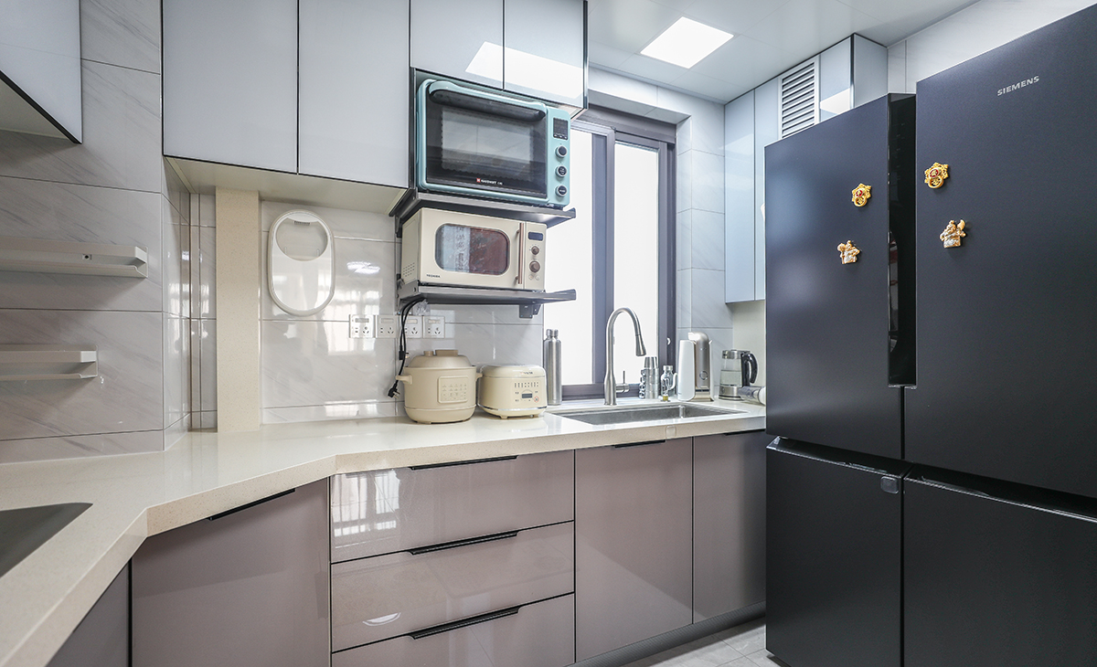 該套房子廚房空間很大，L型三面櫥柜，另外冰箱邊上也有操作臺(tái)，顏色簡潔明快，材質(zhì)要干凈和好打理，白色的吊頂和米色的上櫥柜、深色的下櫥柜相呼應(yīng)，石英石臺(tái)面干凈、好打理，各種廚房電器全部配齊，滿足主人制作各種美食的需求。