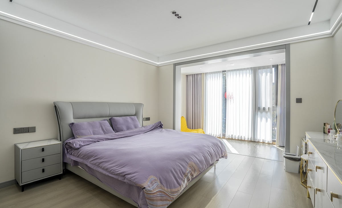  主臥空間很大，頂面有主光源，有輔助光線和氛圍燈，把空間烘托的很浪漫、很溫馨，原木色地板搭配可可蛋奶墻面涂料，灰色的門套和灰色的床相呼應(yīng)，加上紫
