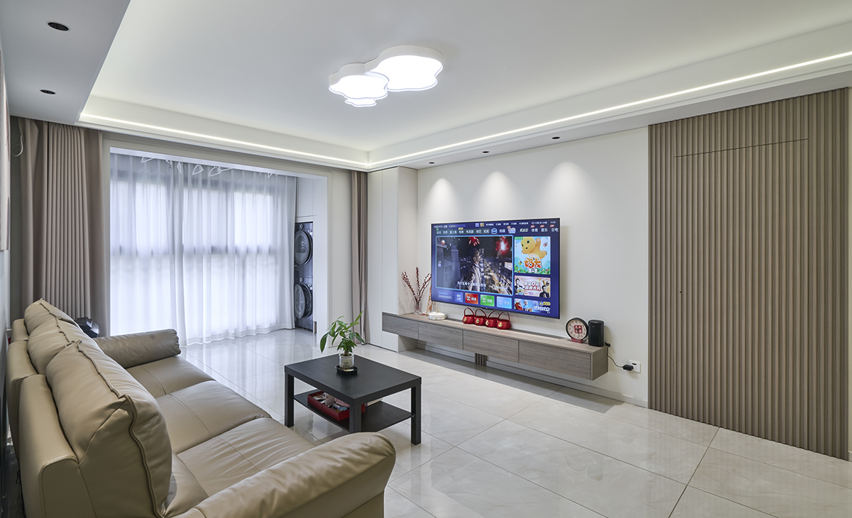 客廳開間很大，電視墻用木格柵作為主臥隱形門，I型的懸空電視柜，咖色皮質沙發立刻檔次提高很多，放一組三人妃沙發，客廳空間也很寬闊。頂面中央空調，吊頂造型上采用雙眼皮吊頂。地面采用暖灰色地磚。造型和色調上無不給人以簡潔、大氣、時尚的感覺。