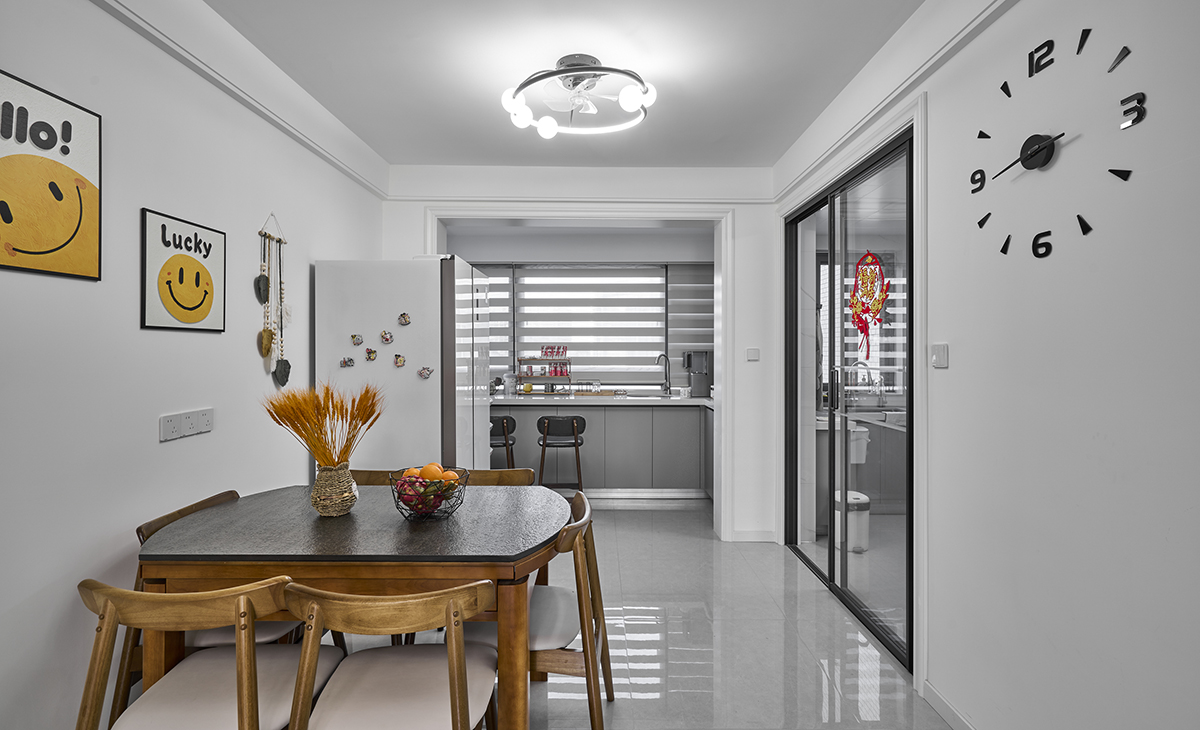 餐廳空間比較方正，餐廳和北陽臺連成一體，灰色的櫥柜和白色的臺面，臺面上配置茶水和裝飾品。作為吧臺使用，兩把高腳凳，。左側是餐廳區域，有一張木質餐桌和四把配套的椅子，桌子上擺放著一個插有干草的花瓶和一盤水果，增添了溫馨的氛圍。墻上掛著兩幅帶有笑臉圖案的裝飾畫，分別寫著“Hello!”和“Lucky”，給空間帶來了活潑的感覺。整個區域的地板是光滑的灰色瓷磚，天花板上有一個現代風格的吊燈，提供充足的照明。這個設計既時尚又實用，適合現代家庭的生活方式。眼皮圍邊的造型，玄關處的花瓶把空間點綴地恰到好處。