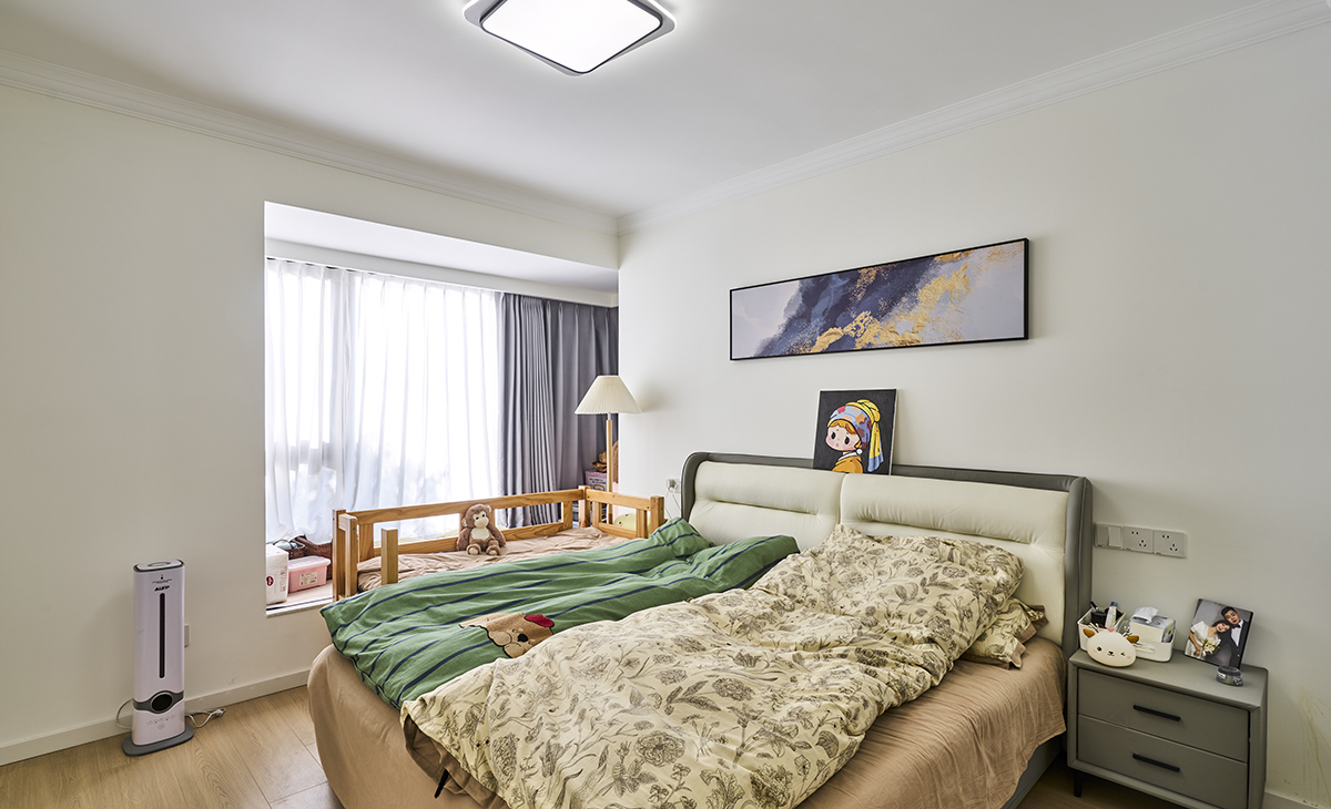 主臥的整體色調以柔和的中性色為主，營造出一種寧靜舒適的氛圍。
床頭靠墊為灰色，與整體色調相協調。
床的右側有一個深灰色的床頭柜，上面擺放了一些個人物品，如相框、護膚品和小擺件，體現了主人的生活氣息。
床頭上方掛有一幅抽象風格的畫作，色彩豐富，為房間增添了藝術感。
畫作下方還有一幅小的裝飾畫，畫中有一個戴著頭飾的女孩形象，增加了房間的趣味性。
房間有一扇大窗戶，窗簾采用了雙層設計，內層是白色的紗簾，外層是深灰色的遮光簾，既能保證光線的柔和進入，又能在需要時提供良好的遮光效果。
窗戶旁邊放置了一張木質嬰兒床，床上有一些玩具，是為小寶設計的，體現了溫馨和關愛的氛圍。
 床頭柜上方有一盞簡約風格的壁燈，提供了夜間照明，同時也作為房間的裝飾元素。
 
整個主臥設計簡潔而不失溫馨，注重細節和功能性，適合現代家庭的居住需求。