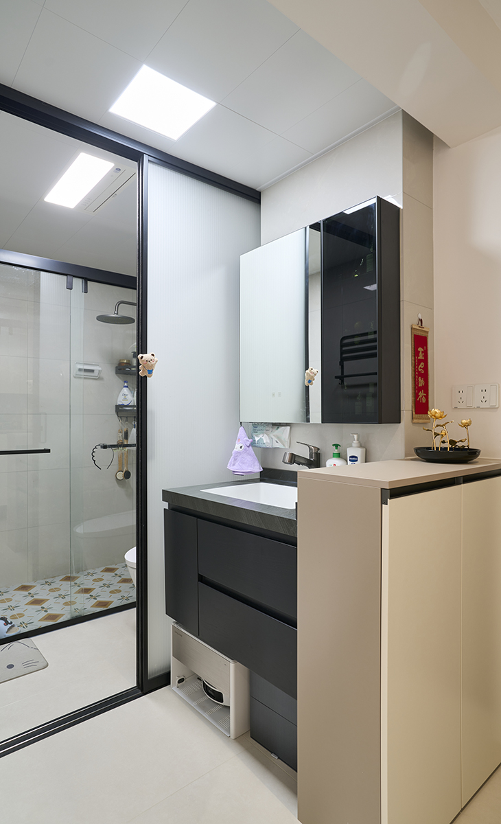 衛(wèi)生間采用干濕分離的布置，淺灰色墻磚配深灰色色浴室柜和黑色邊框長(zhǎng)虹玻璃移門(mén)，顏色明暗有序，從深到淺，色調(diào)很豐富，掛墻臺(tái)盆柜，墻排水設(shè)計(jì)，下方又設(shè)計(jì)一個(gè)掃地機(jī)器人，顏色協(xié)調(diào)統(tǒng)一，主人很滿(mǎn)意。  
