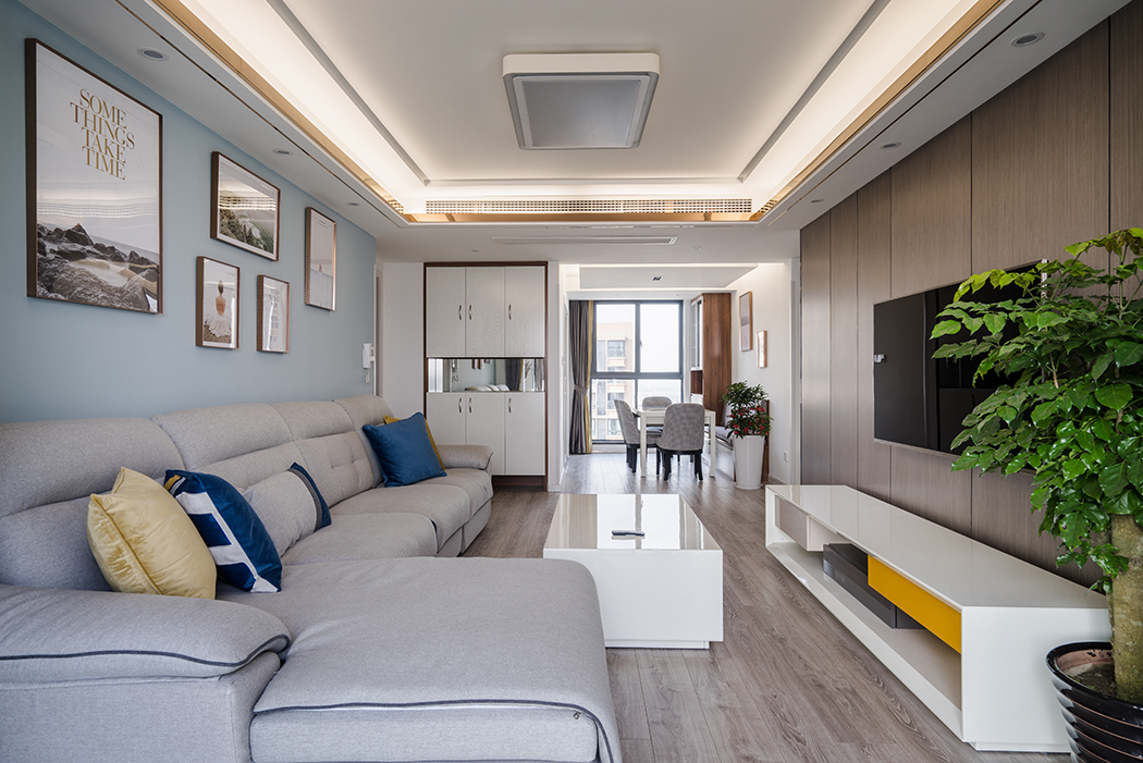 而本案空間采用原木色系的色調，主空間靜謐藍的沙發背景和紋理清晰的木飾面電視背景相互呼應。同時靜謐藍的背景襯托了淡灰色輕奢的布藝家具及軟裝畫，讓空間不在拘謹，在陶瓷白的茶幾的對比下產生色彩的碰撞，造型簡潔的家具設計透露出幾分優雅怡然的空間氣質。
