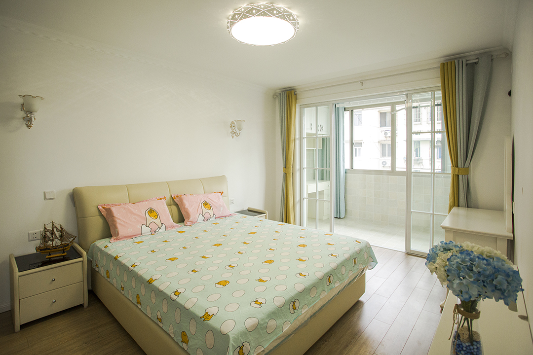 床頭柜上方的壁燈和正中間的吸頂燈各自分工又互相映襯，照亮奢華大氣的軟包雙人床，靜謐安寧。藍白相間的花瓶和精致的梳妝臺似乎都在訴說著主人的幸福和浪漫。
