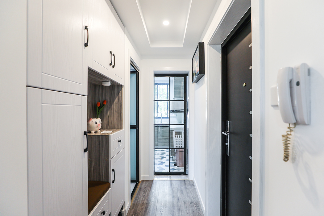 將鞋柜與其他收納空間整合至一側墻面，視線前方廚房門運用簡約幾何形元素，光線自然穿過狹小過道，使小屋充滿溫馨感。
