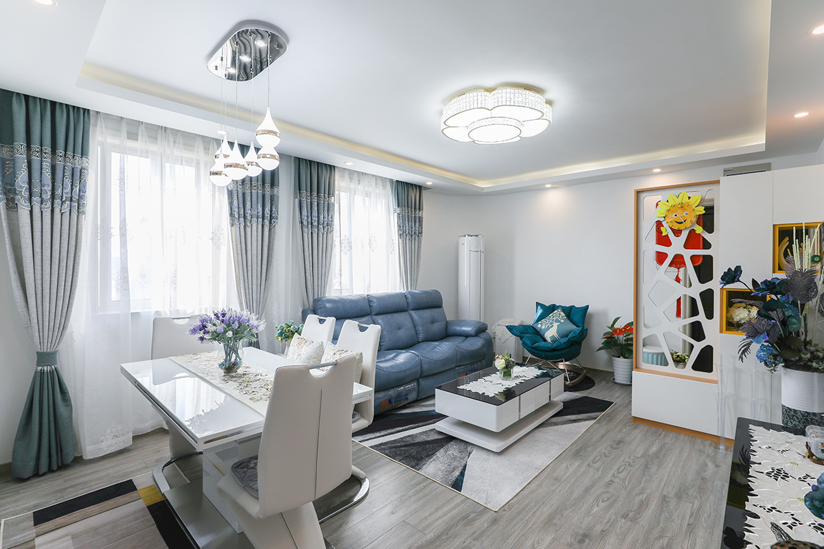 客餐廳頂面平頂，四周做造型頂，配以燈槽及筒燈作為氛圍燈。墻頂面白色乳膠漆干凈簡潔。地面鋪裝深色地板，色彩重心平穩。淺藍色窗簾及藍色沙發相近色配色顯得空間既清爽又有層次感。
