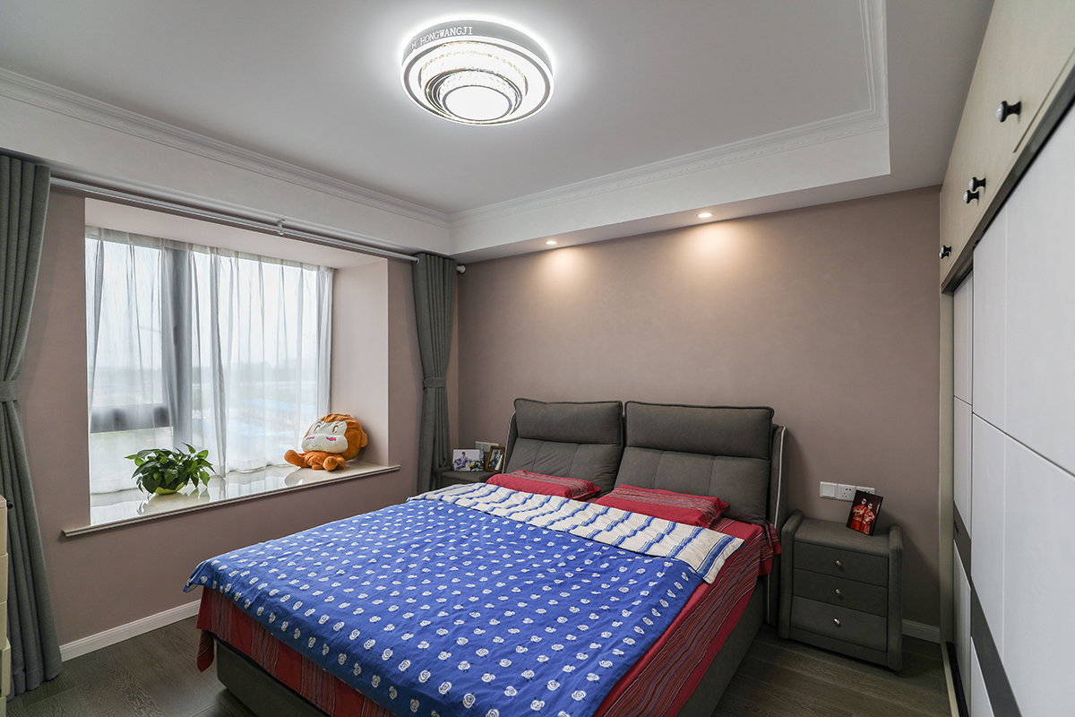 相比于轟轟烈烈的愛情，或許你更傾向于細水長流的相遇相知！主臥的設計簡約大方，深色布藝大床搭配白藍色床單，輔以實木床頭柜、灰白色窗簾和小筒燈做點綴，整個空間溫馨舒適。
