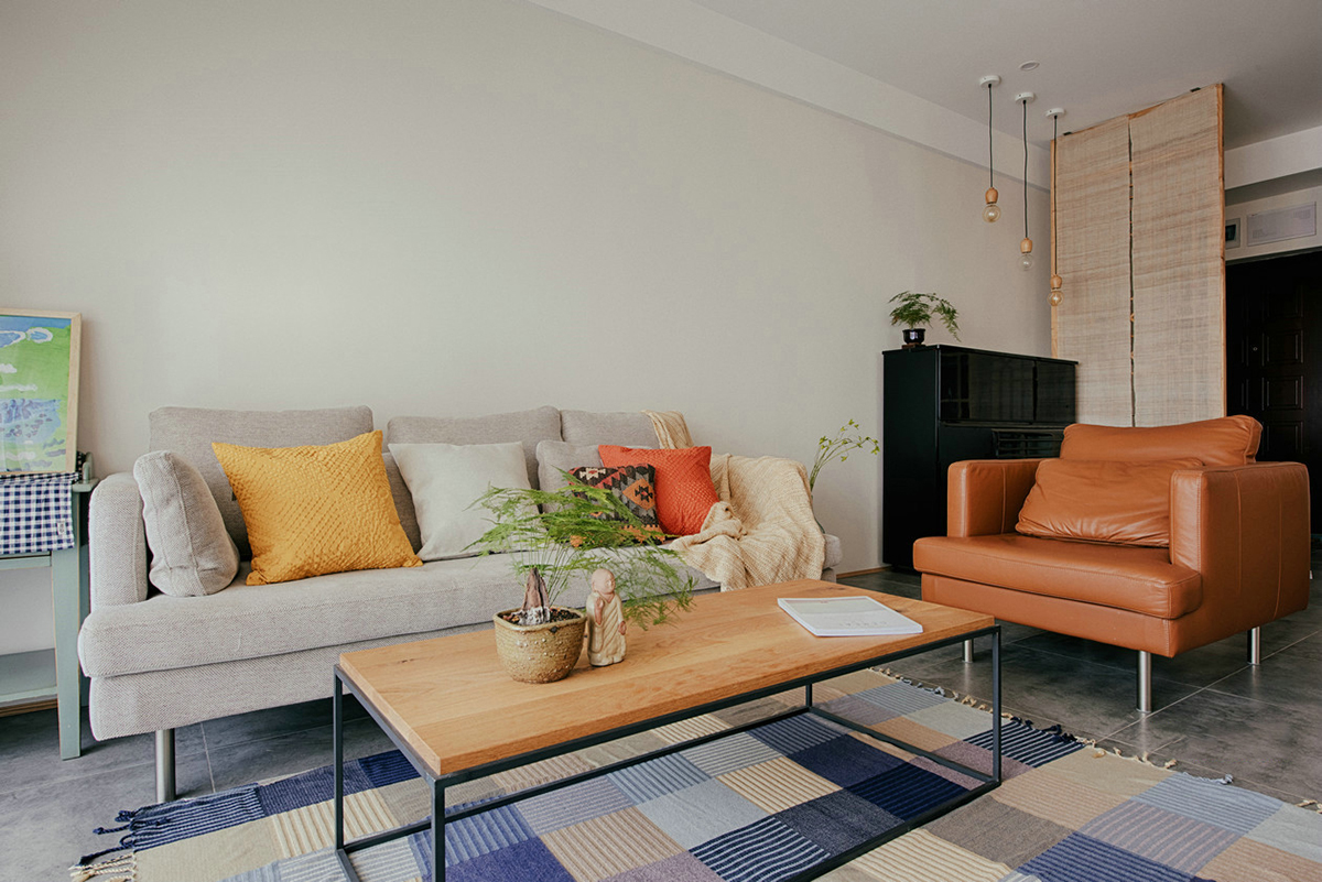 客廳空間開闊明亮。利用淺色紗幔窗簾優化自然光的吸采，讓空間擁有足夠的恬靜感。業主對日式風格很喜歡，客廳的茶幾是業主在日本旅游時帶回國?；疑牟妓嚿嘲l，沉穩大氣，搭配自然紋理的原木茶幾和座椅，讓濃郁的日式風情填滿整個空間。柔軟舒適的沙發，隨意擺放著幾個抱枕，再增加一張橙色的真皮單人椅，無形之中提升了空間的質感。
